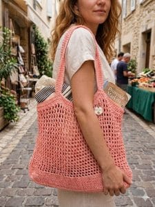 Fiatal nő mediterrán stílusú, macskaköves utcán sétál, a vállán egy BeLuna Műhely korall színű horgolt táskával, amiben egy 'Provence Markets' feliratú könyv van.